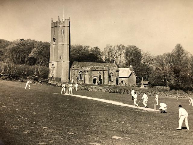 A old photo of a church and a cricket pitch. There is a game of cricket in progress.
